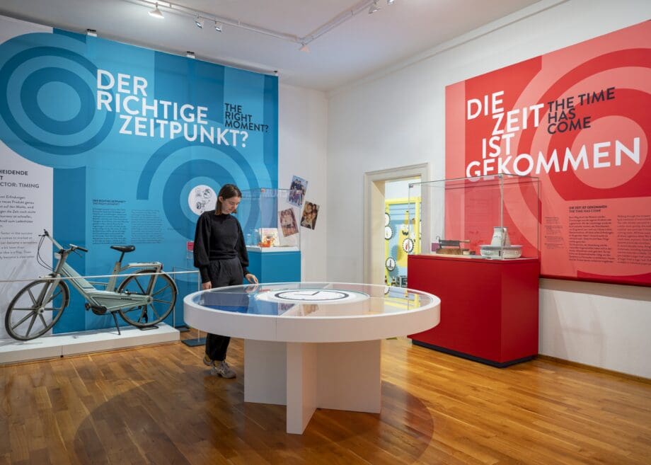 Austellungsraum mit Objekten, Vitrinen und einer Person, die sich Objekte in einer Vitrine ansieht.